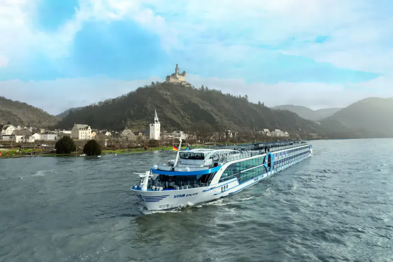 White luxury river cruise ship sails on the Rhine past Marksburg Castle on hill, with village and mountains under blue sky.
