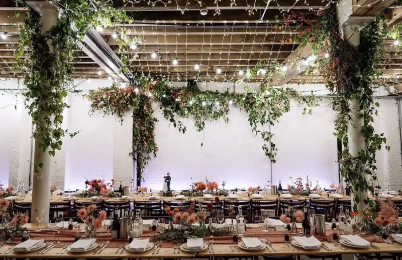 Elegant wedding reception table in industrial venue with ivy vines, fairy lights, and orange floral arrangements.