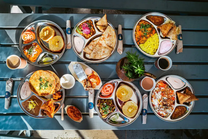 Overhead view of multiple thali plates with Indian dishes: curries, rice, naan, veggies on wooden table.