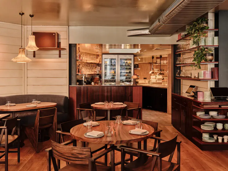 Cozy interior of AngloThai restaurant with wooden tables set with glasses and napkins, bar, and potted plants.