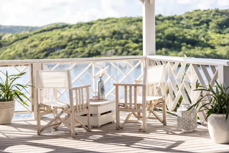 Two white director's chairs and small table with vase on sunny wooden balcony overlooking lush green hills and lake at Rokuni, Antigua.