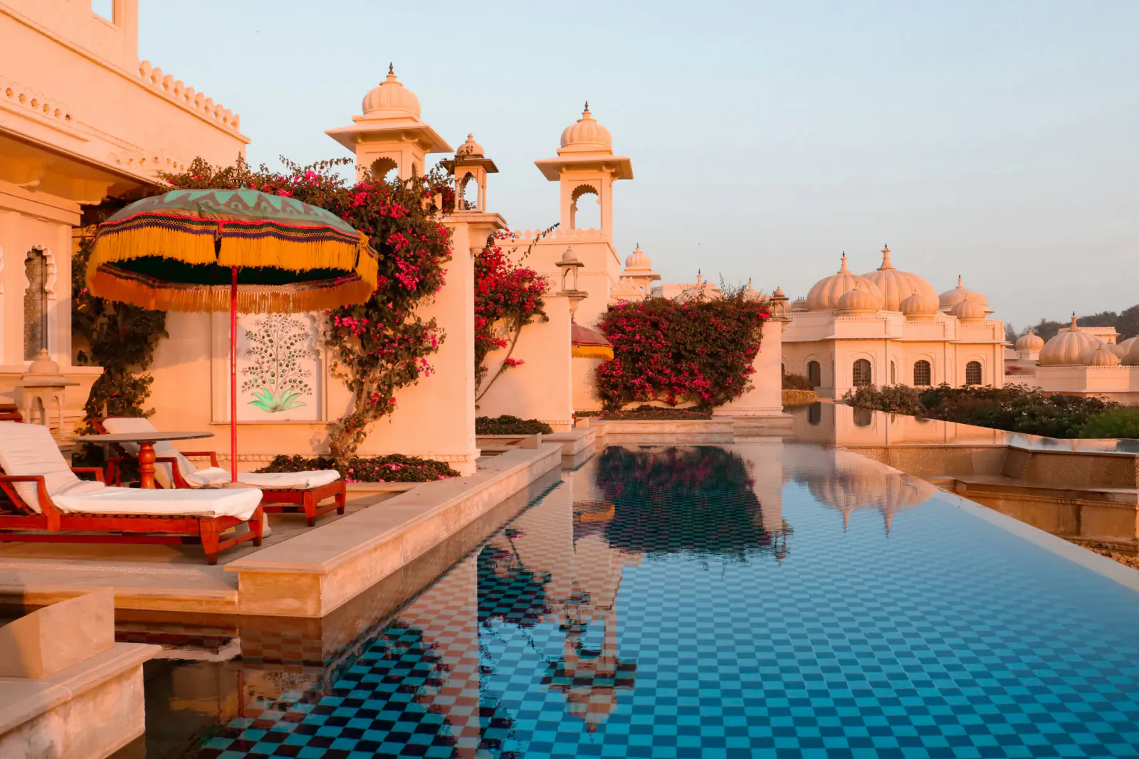 Luxury Indian palace rooftop infinity pool at dusk with orange umbrella, loungers, bougainvillea, and domes.