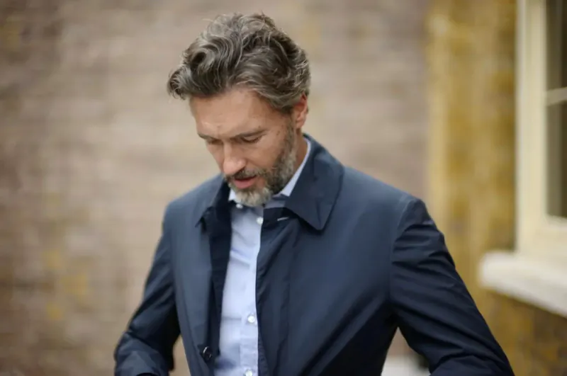 Man with gray hair and beard in navy bespoke jacket and white shirt, thoughtful pose against brick wall.