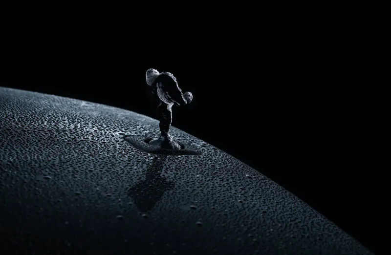 Rolls-Royce Spirit of Ecstasy figurine on Black Badge Ghost hood ornament base atop a dark, wet spherical surface against black space.