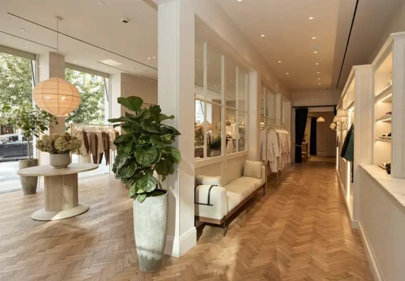 Varley SoHo flagship store interior: white hallway with clothing racks, plants, round table, and bench amid herringbone floors.