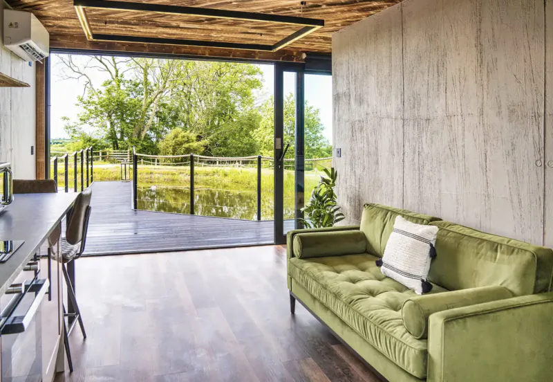 Modern interior of The Boathouse luxury lodge with green velvet sofa, bar stools, and sliding doors to deck overlooking pond and trees.