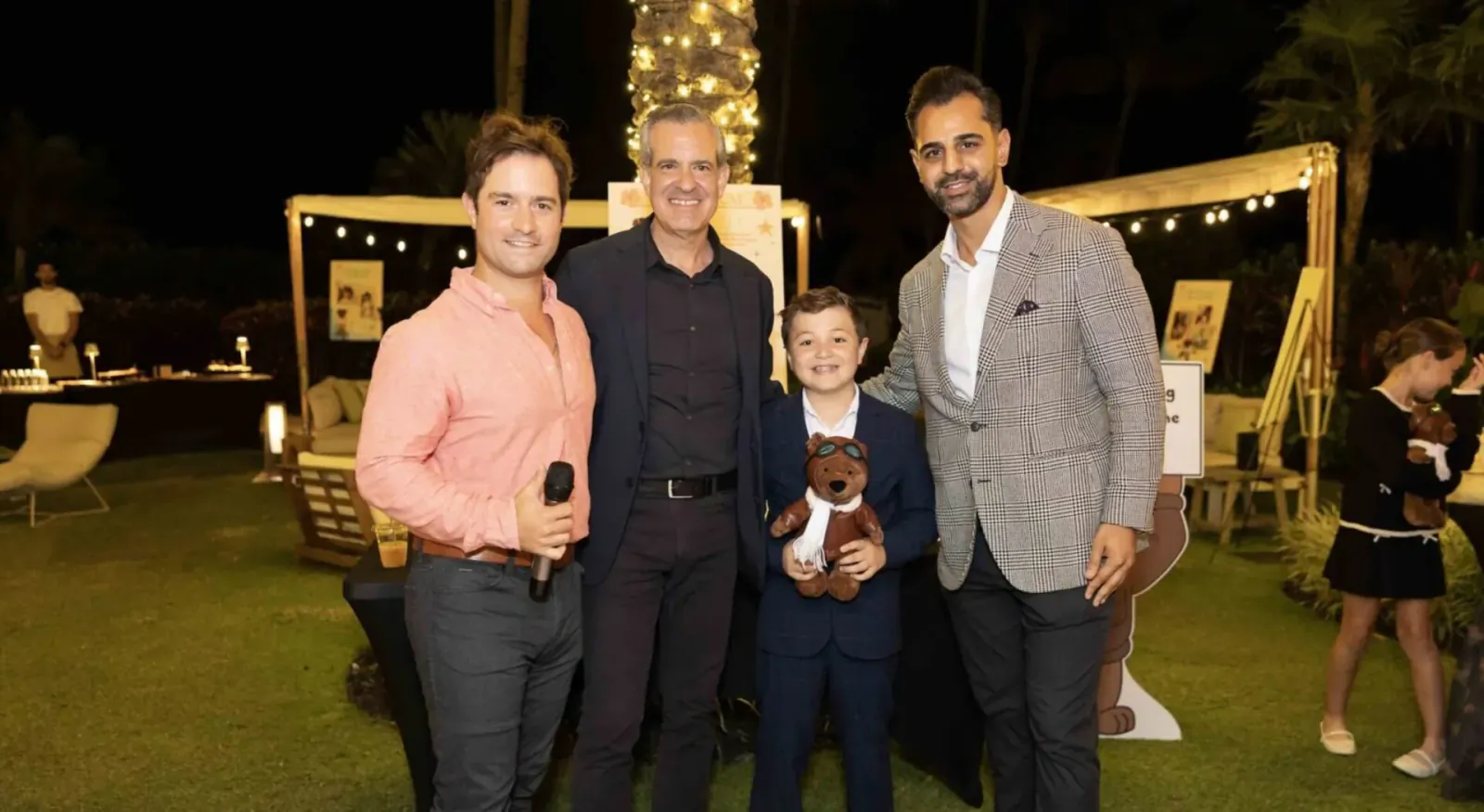 Four men and a young boy in suit holding teddy bear pose smiling at outdoor evening event with golden Christmas tree and palms.
