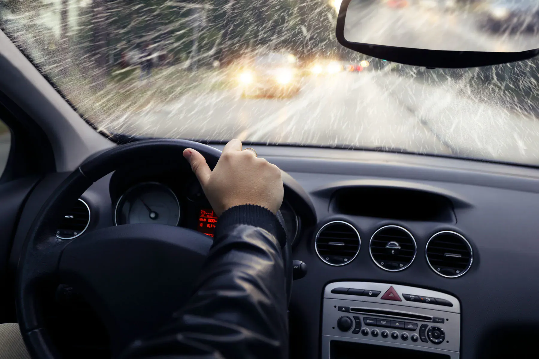 Close-up of driver's hand on steering wheel in rainy car, wet windshield and road ahead.