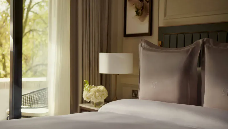 Luxurious hotel bedroom at Carlton Tower Jumeirah, London: plush bed with pillows, lamp, white orchids, large window with autumn trees.