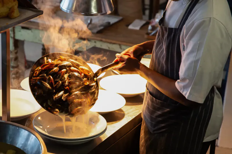 Chef pouring steaming stir-fried prawns from wok onto white plate in busy kitchen at Olympia Café