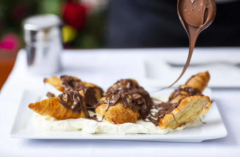 Close-up of croissants on a plate topped with chocolate sauce being poured from a spoon, on white cream, with flowers in background.