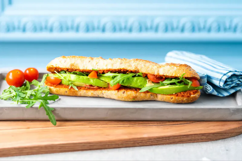 Baguette sandwich with avocado, tomatoes, arugula on slate board, cherry tomatoes, blue napkin, wood backdrop