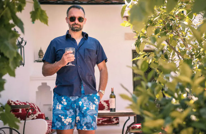 Man in navy shirt and floral swim shorts holds wine glass outdoors by white wall, greenery, table with wine bottle in Marbella.