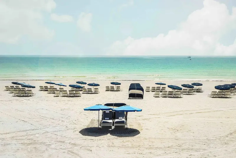 Sandy beach with rows of blue umbrellas and lounge chairs by turquoise sea under partly cloudy sky