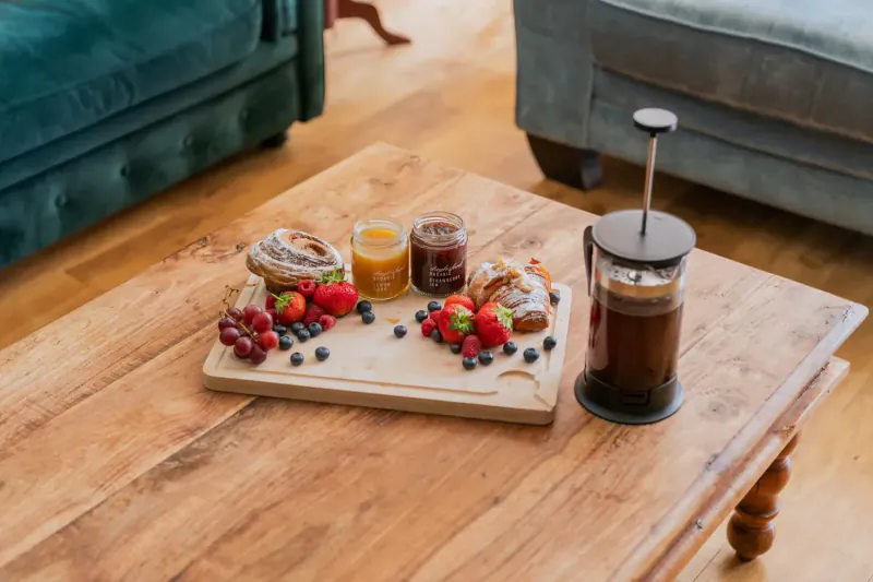 Wooden board on rustic table with pastries, jam jars, berries, and French press coffee, green velvet chairs in background.