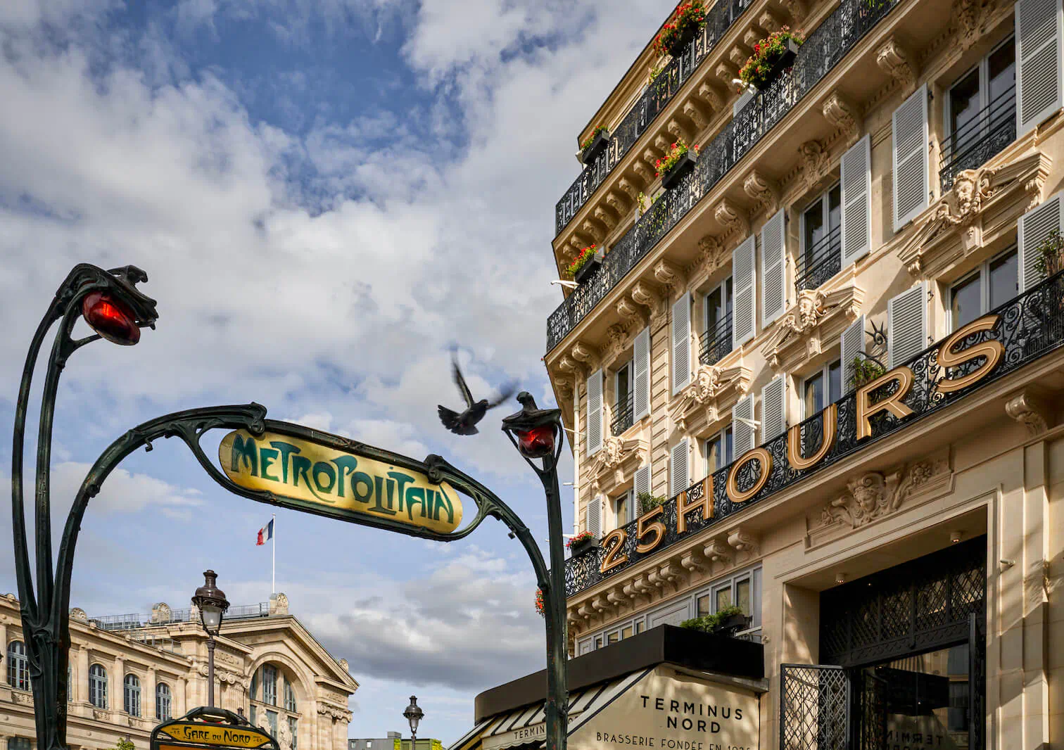 Iconic Paris Métro sign arching over 25Hours hotel entrance with ornate facade, flowers, French flag, and cloudy sky.