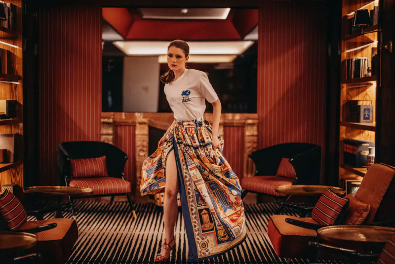 Woman in white graphic tee and flowing colorful skirt poses dynamically in VINHA Boutique Hotel library lounge with red walls and bookshelves.