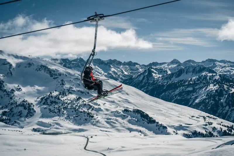 Skier in red jacket on chairlift skis over snowy Val d'Aran mountains under blue sky with clouds.