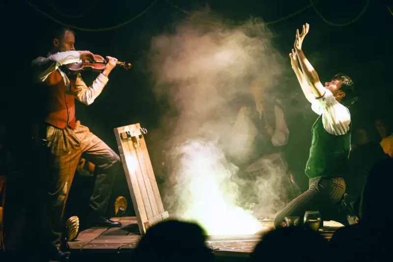 Two performers on stage: one plays violin in vest, other raises arms by fire-lit board amid smoke at Christmas show.