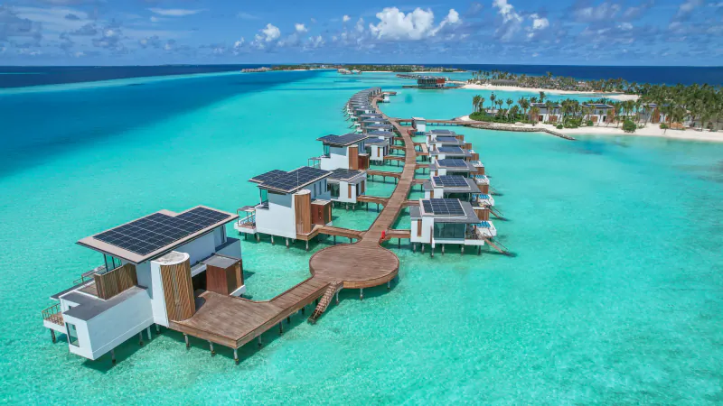 Aerial view of modern overwater villas with solar panels on wooden walkways in turquoise lagoon at Crossroads Maldives