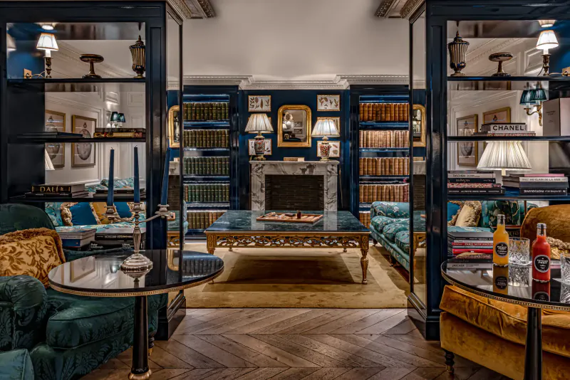 Elegant Parisian library lounge at Relais Christine with navy bookshelves, marble fireplace, emerald velvet sofas, and orange drinks on tables.