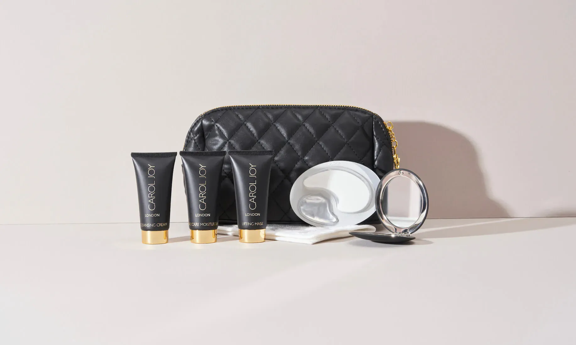 Carol Joy skincare set: four black tubes with gold caps, white dish with cloth, round mirror, in black quilted pouch