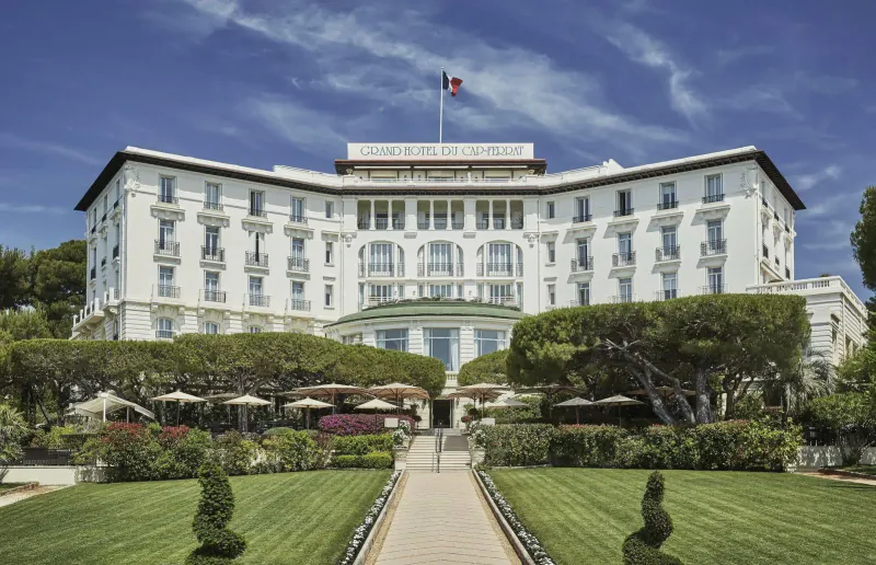 Grand white luxury hotel facade with French flag, manicured gardens, pine trees, and pathway under blue sky