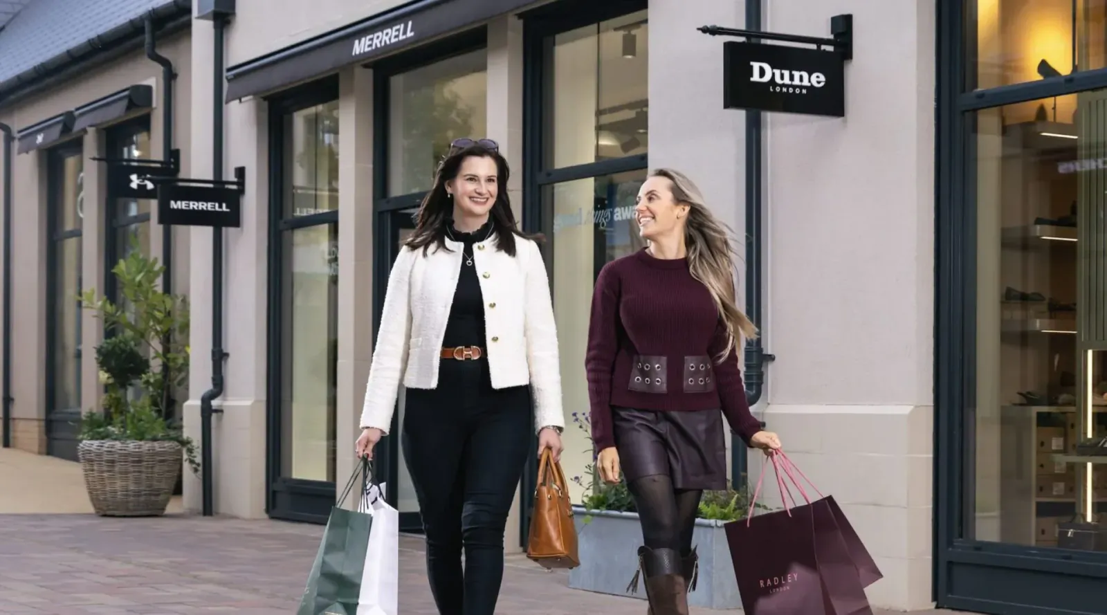 Two stylish women in white blazer and maroon sweater carry Dune shopping bags, walking outside Dune store.