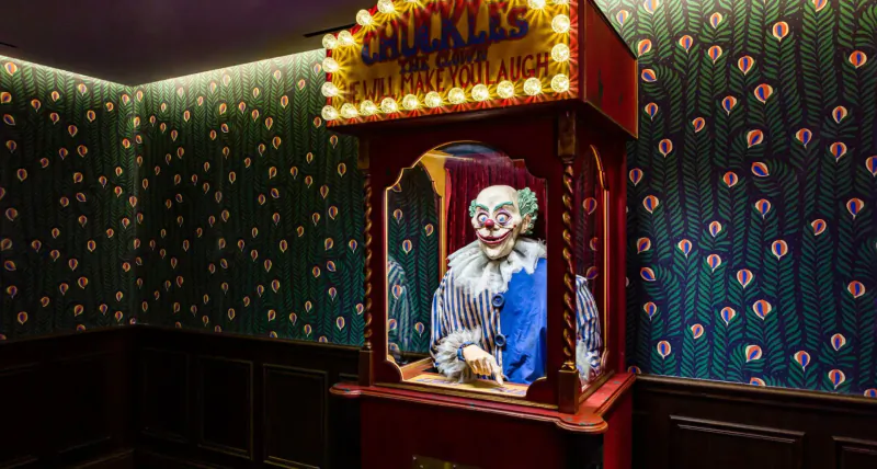 Chuckles Fortune Teller clown puppet in ornate red booth with neon sign, against floral green patterned walls in Monarch Theatre gallery.