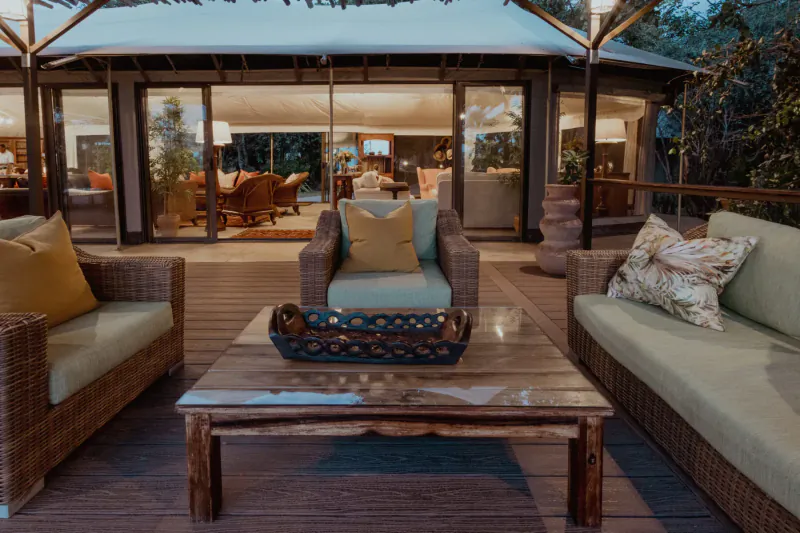 Luxury canvas lodge deck at The Elephant Camp with cushioned wicker furniture around a wooden coffee table, evening jungle setting.