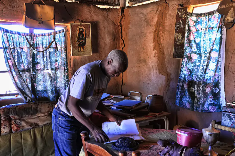 Bald man in t-shirt and jeans bent over wooden table reading papers in rustic adobe room with blue curtains and icon