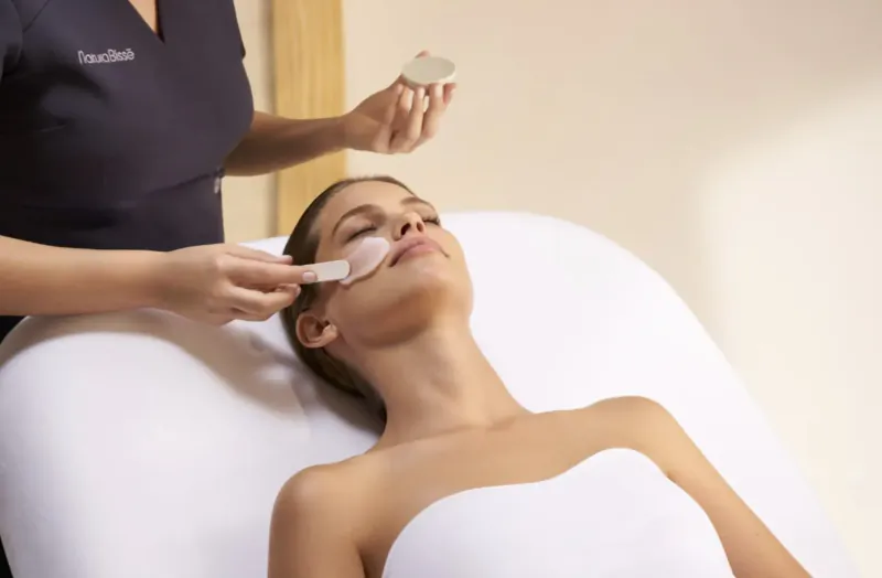 Spa therapist applies white facial mask with spatula to relaxed woman reclining on treatment bed