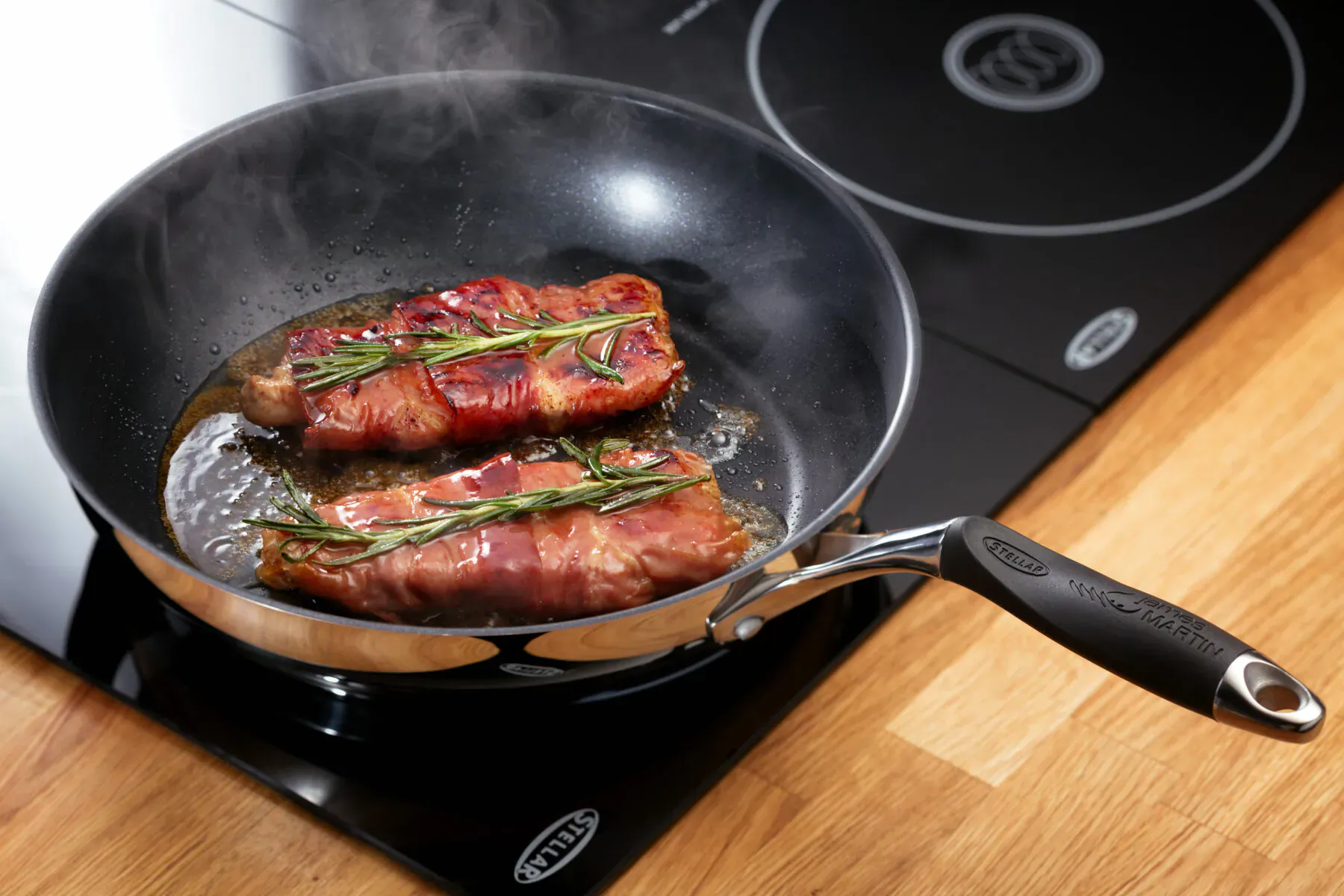 Two rosemary-wrapped bacon fillets sizzling in a Tefal frying pan on an induction hob.
