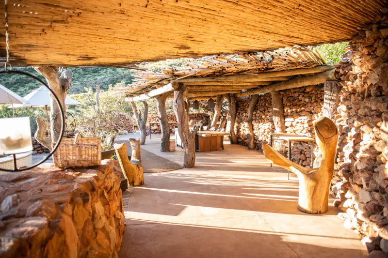 Rustic thatched veranda at Tarkuni, Tswalu, with log pillars, stone walls, wooden furniture, and mountain backdrop.