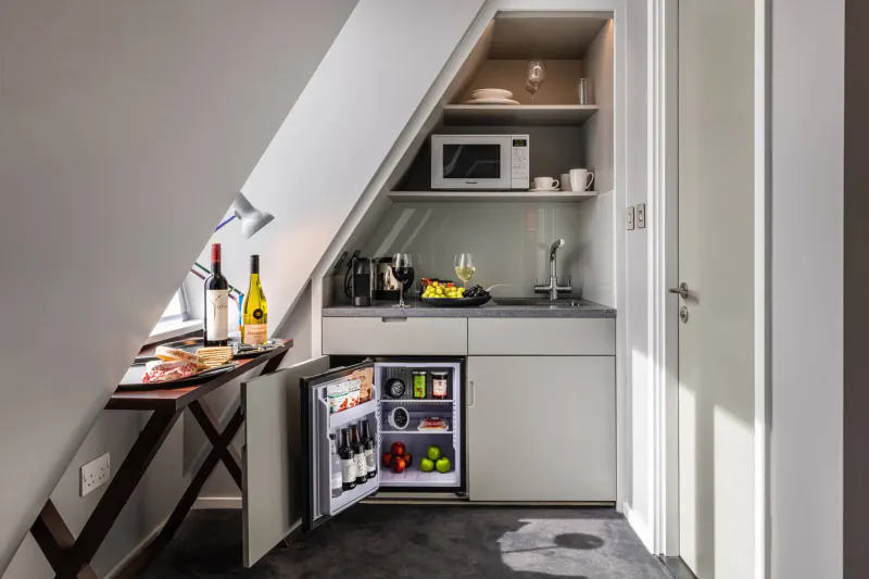 Compact attic kitchenette in London hotel with sloped ceiling, mini fridge, microwave, wine bottles, coffee maker, and counter snacks.