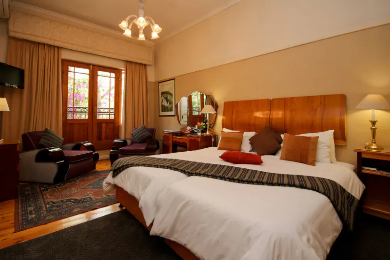 Elegant hotel room at Montagu Country Hotel with wooden headboard bed, armchairs, balcony, and chandelier.