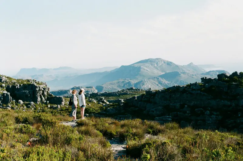 Couple in white stands on grassy mountain path overlooking Cape Town's misty Twelve Apostles peaks