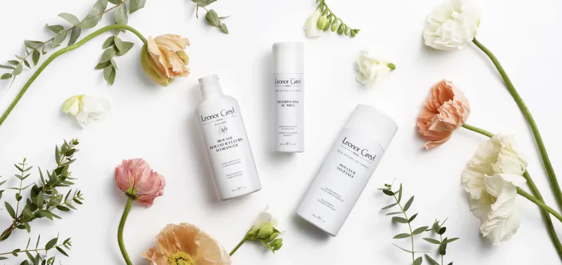 Leonor Greyl hair care products: white bottles and jars arranged with white, pink, and orange flowers on white background