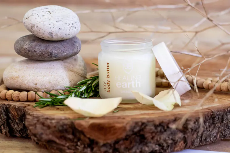 Open jar of Healing Earth Body Butter on wooden surface with stacked stones, rosemary sprigs, and petals