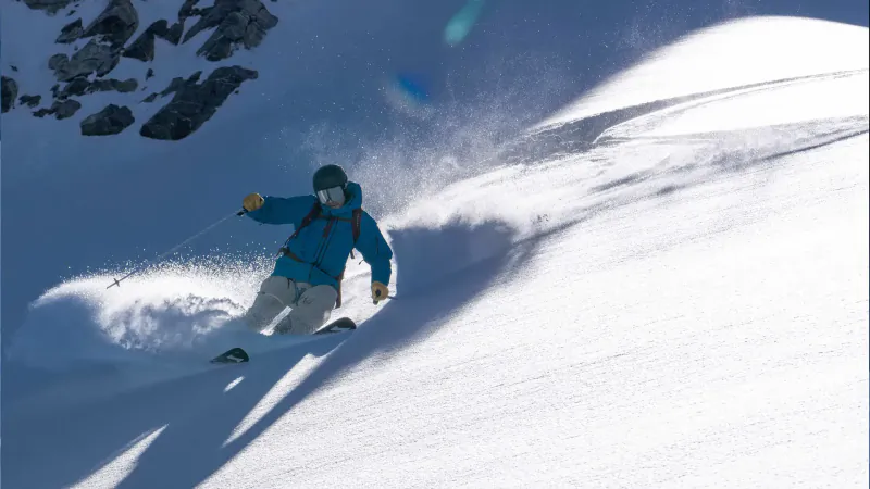 Skier in blue jacket carves snowy slope, kicking up powder amid rocks and sunlight.