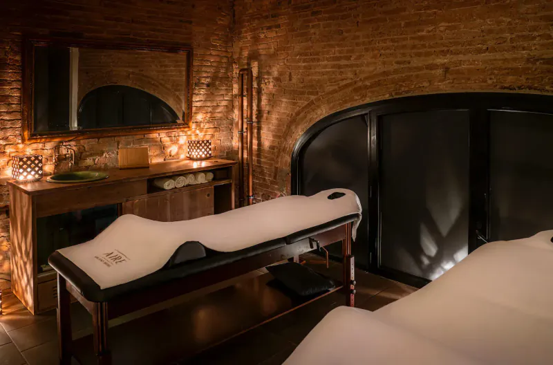 Luxury candle-lit spa room at Aire Ancient Baths with two white-sheeted massage tables, brick walls, mirror, and wooden cabinet.