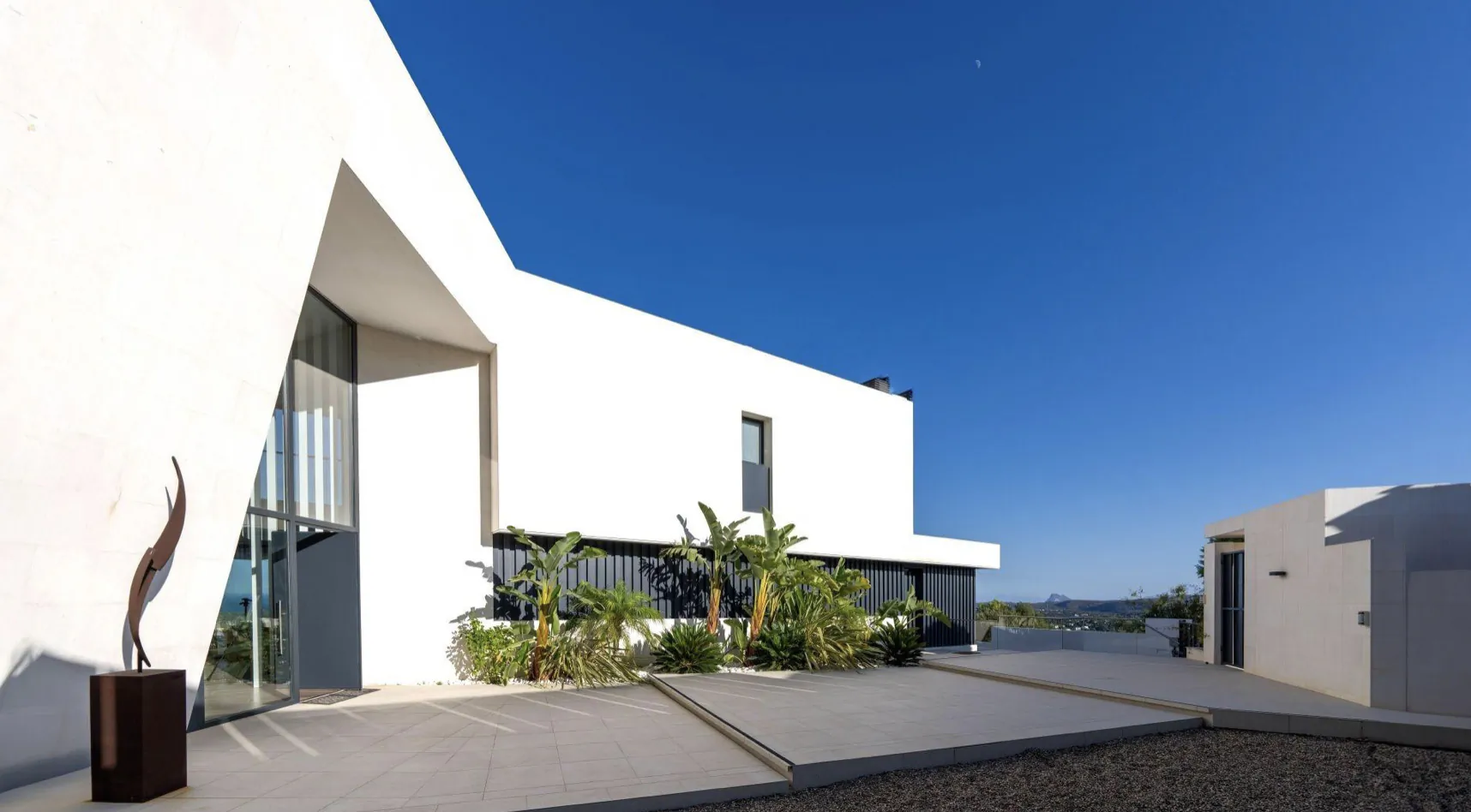 Modern white angular house with abstract sculpture, palms, and pool under clear blue sky