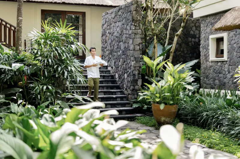 Man in white shirt stands on stone steps amid lush tropical plants at Kayumanis Ubud Bali resort.