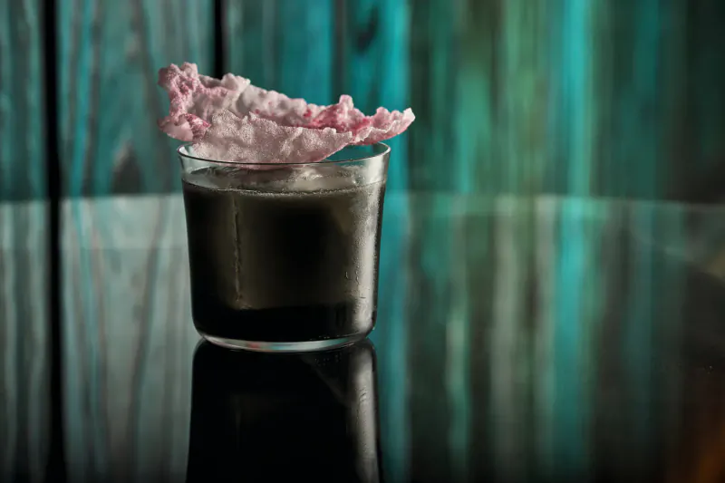 Cocktail glass with dark liquid and pink fluffy garnish on wooden table, teal background blur, Halloween event vibe.