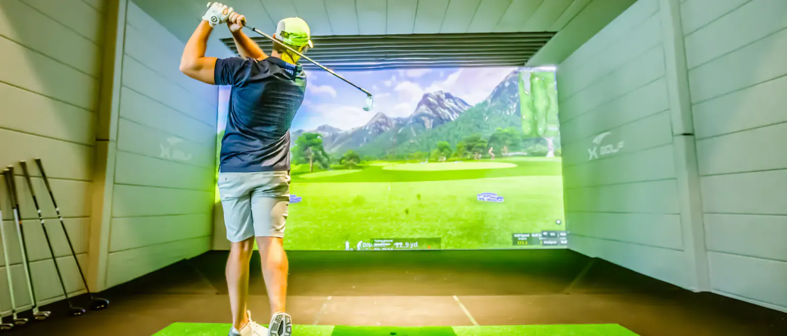 Man in black shirt and shorts swings golf club on simulator screen showing Hideaway Beach Resort golf course with mountains.
