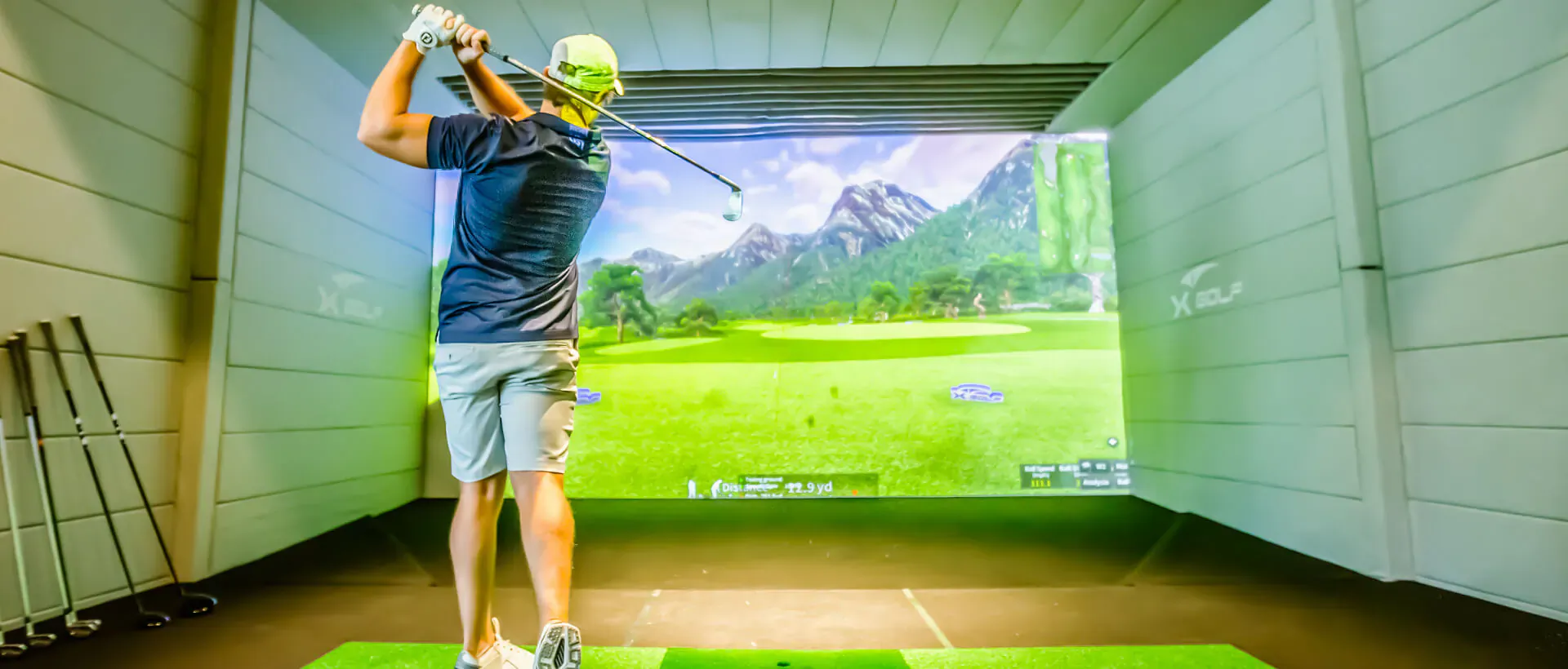 Man in black shirt and shorts swings golf club on simulator screen showing Hideaway Beach Resort golf course with mountains.
