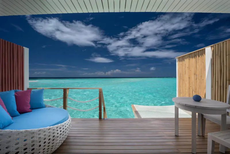 Cora Cora Maldives overwater villa deck with blue cushions, table, and turquoise ocean under cloudy blue sky
