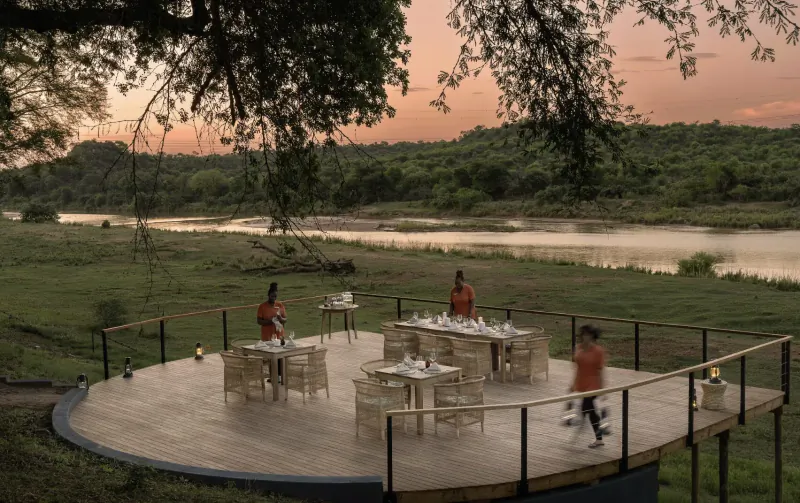 Aerial view of women in red uniforms setting tables on a circular wooden deck overlooking river at sunset in wilderness.