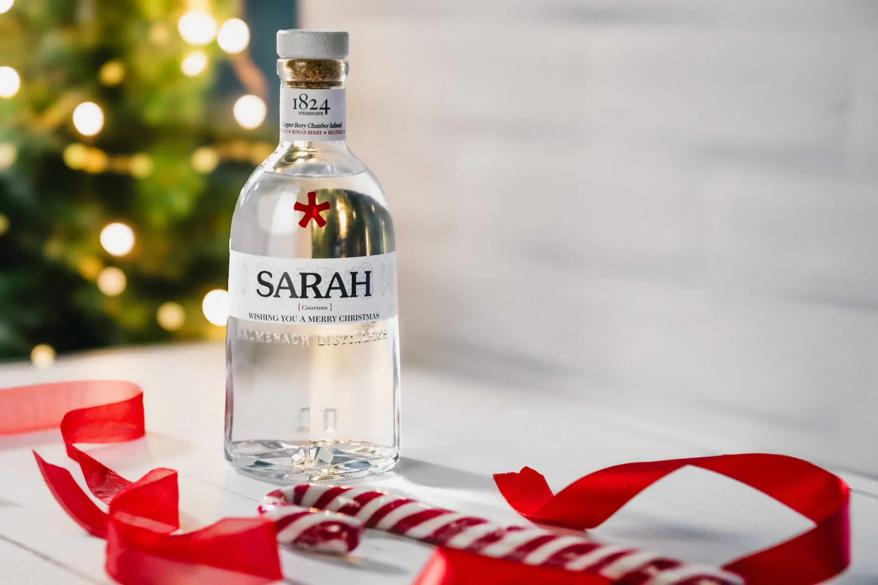 Sarah gin bottle with red cross label on white surface, red ribbon, festive Christmas tree lights bokeh background