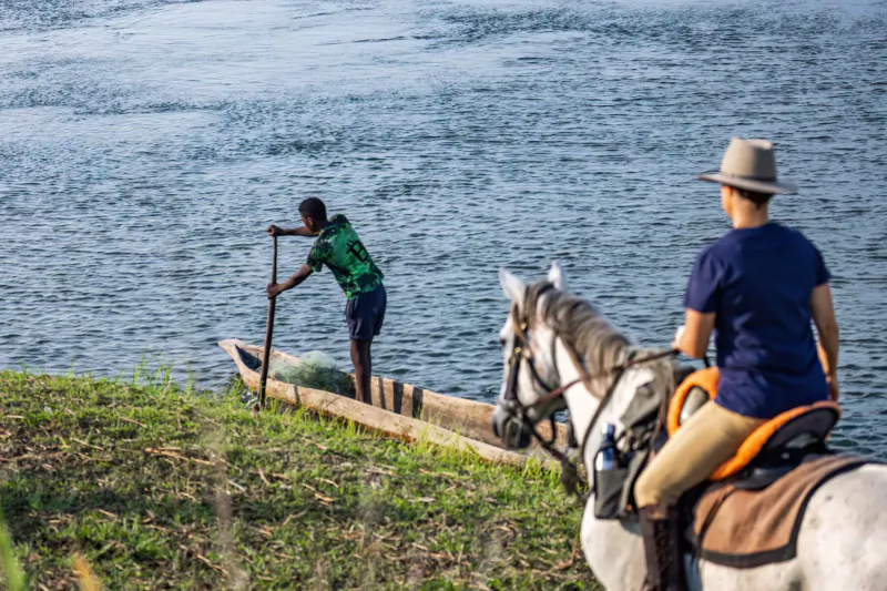Man in green shirt paddling dugout canoe on riverbank as rider on white horse with orange saddle watches, Zambian safari scene.